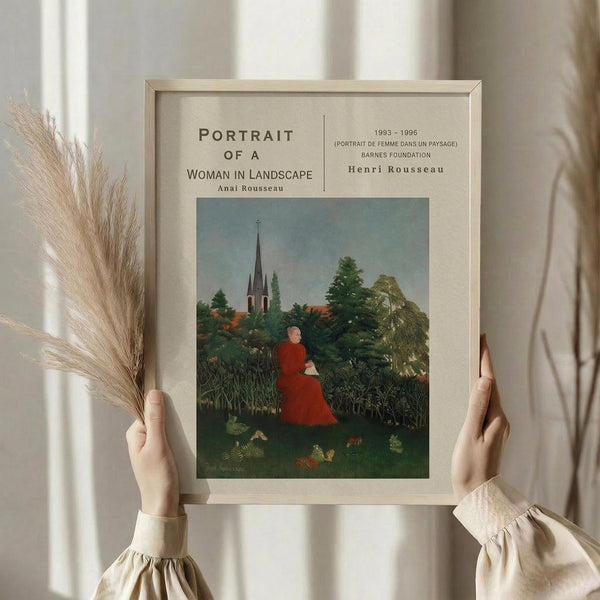 May include: Person in cream ruffle-sleeve top holding framed art print showing woman in red dress sitting in lush green landscape with church spires in background. Natural wood frame, cream matting. Dried wheat stalks visible on left. Soft natural lighting against white curtained window.