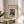 May include: Framed art print displaying a Japanese woodblock-style winter scene with snow-covered cedar trees, small figures walking on a snowy path, and traditional buildings. The artwork features muted blues, browns, and whites against a cream background. Shown in a thin wooden frame on a round light wood side table next to a beige upholstered chair, with plant leaves visible and soft window light creating shadows on white walls.