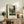 May include: Framed art print of Henri Rousseau's "Toll Gate" painting displayed on rustic wooden sideboard in warm neutral living room. Print shows pastoral scene with figures near gate, surrounded by lush green trees and countryside. Light wood frame with white matting. Scene includes cream upholstered chair, fern plant, ceramic vase, and stacked books creating cozy home decor styling.