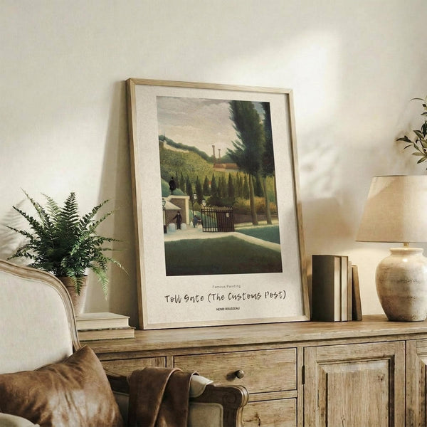 May include: Framed art print of Henri Rousseau's "Toll Gate" painting displayed on rustic wooden sideboard in warm neutral living room. Print shows pastoral scene with figures near gate, surrounded by lush green trees and countryside. Light wood frame with white matting. Scene includes cream upholstered chair, fern plant, ceramic vase, and stacked books creating cozy home decor styling.
