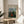 May include: Framed art print on light wood round table beside beige upholstered chair. Print shows woman in red dress sitting in lush green landscape with church spire visible through trees. Poster has cream background with black text above colorful naive-style painting. Soft natural lighting creates shadows from nearby plant leaves on white wall.