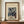May include: Framed vintage Japanese woodblock print showing snow-covered cedar trees and small figures walking through a winter forest scene. The artwork features muted blues, browns, and whites with falling snow. Displayed in a dark wooden frame on a light wood sideboard next to a cream ceramic vase and stacked books against white paneled walls.