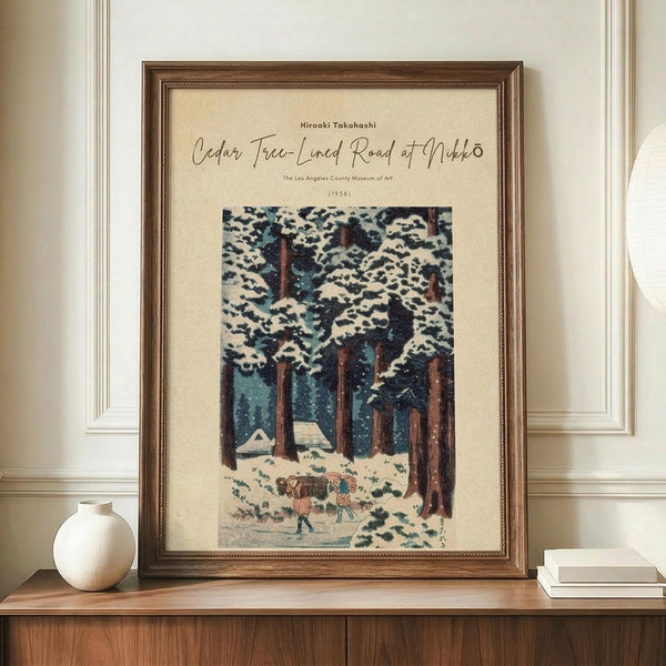 May include: Framed vintage Japanese woodblock print showing snow-covered cedar trees and small figures walking through a winter forest scene. The artwork features muted blues, browns, and whites with falling snow. Displayed in a dark wooden frame on a light wood sideboard next to a cream ceramic vase and stacked books against white paneled walls.