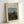 May include: Framed art print showing a woman in red dress sitting in lush green landscape with church spire in background. Light wood frame with white matting displays reproduction of naive-style painting with detailed foliage and pastoral scene. Print leaning against white wall on light surface, showing museum-quality presentation with artist and date information at top.