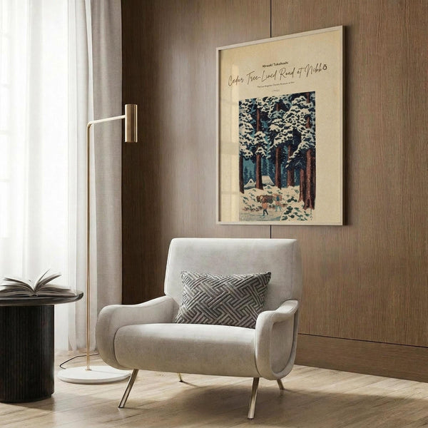 May include: A framed vintage Japanese woodblock print showing a snow-covered forest path with tall cedar trees and small figures walking through winter landscape. The cream-colored poster features blue and brown tones with white snow details. Displayed on wood-paneled wall above a beige upholstered armchair with geometric patterned pillow, next to a brass floor lamp and dark side table with open book in modern interior setting.