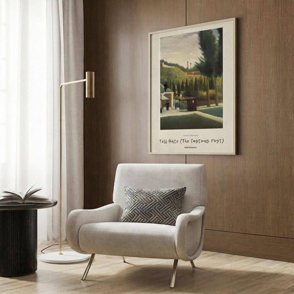 May include: Framed art print of pastoral landscape with toll gate, trees, and countryside scene in green and earth tones, displayed on wood-paneled wall above cream upholstered armchair with geometric patterned pillow, brass floor lamp and side table with open book nearby in modern living room setting.