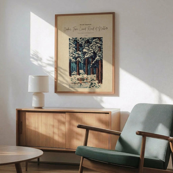 May include: Framed vintage Japanese woodblock print showing snow-covered cedar trees with small figures walking on a path below, displayed above a mid-century modern wooden credenza with ceramic table lamp. Green upholstered armchair with wooden frame sits nearby on hardwood floor with natural sunlight casting shadows across white wall.