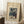 May include: Framed art print leaning against white wall corner on textured cream rug. Shows winter forest scene with snow-covered cedar trees, small figures walking on snowy path. Cream matting in light wood frame. Coffee cup on wooden block nearby, soft natural lighting creates window shadows on wall.