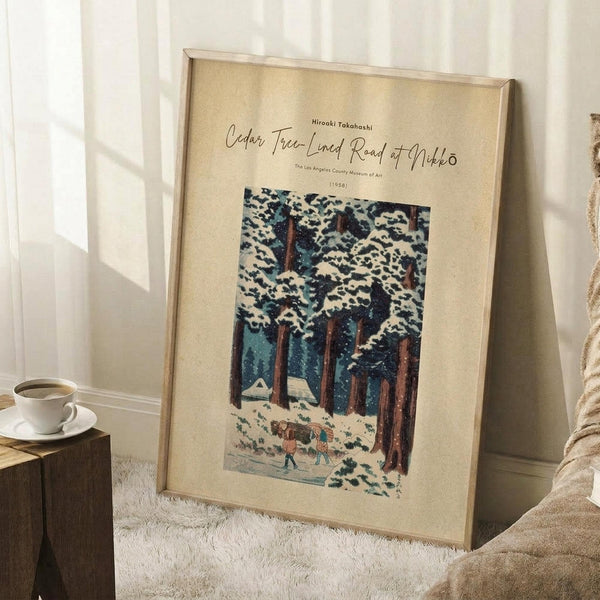 May include: Framed art print leaning against white wall corner on textured cream rug. Shows winter forest scene with snow-covered cedar trees, small figures walking on snowy path. Cream matting in light wood frame. Coffee cup on wooden block nearby, soft natural lighting creates window shadows on wall.