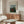 May include: A framed art poster showing a woman in a red dress standing in a lush green landscape with trees and a church spire in the background. The print is displayed above a wooden sideboard in a modern living room with a white curved chair, glass coffee table, and minimalist white table lamp. The poster has a beige border with black text and appears to be in a thin wooden frame against white walls.