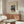 May include: Vintage Japanese woodblock print poster showing snow-covered cedar trees and small figures on a winter path, displayed in wooden frame above mid-century walnut credenza. Room features cream textured chair, glass oval coffee table, white table lamp, and minimalist decor with soft natural lighting.
