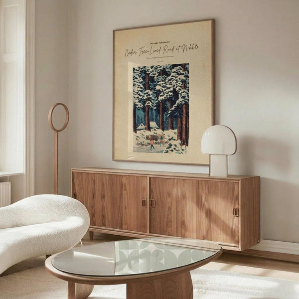 May include: Vintage Japanese woodblock print poster showing snow-covered cedar trees and small figures on a winter path, displayed in wooden frame above mid-century walnut credenza. Room features cream textured chair, glass oval coffee table, white table lamp, and minimalist decor with soft natural lighting.