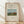 May include: Framed vintage art poster featuring Henri Rousseau's pastoral painting of a cow grazing in a green field with large haystacks, trees, and church spire in background. Cream-colored poster with black text reading "Scene in Bagneux on the Outskirts of Paris 1909" and artist signature. Natural wood frame leaning against white wall on light wooden floor with soft shadows.