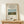May include: Framed art print displaying a pastoral scene with a cow grazing in green countryside, haystacks, and distant church spire under blue sky. Cream matted poster in light wood frame sits on wooden bench beside small wrapped gift with botanical pattern and twine bow against white wall.