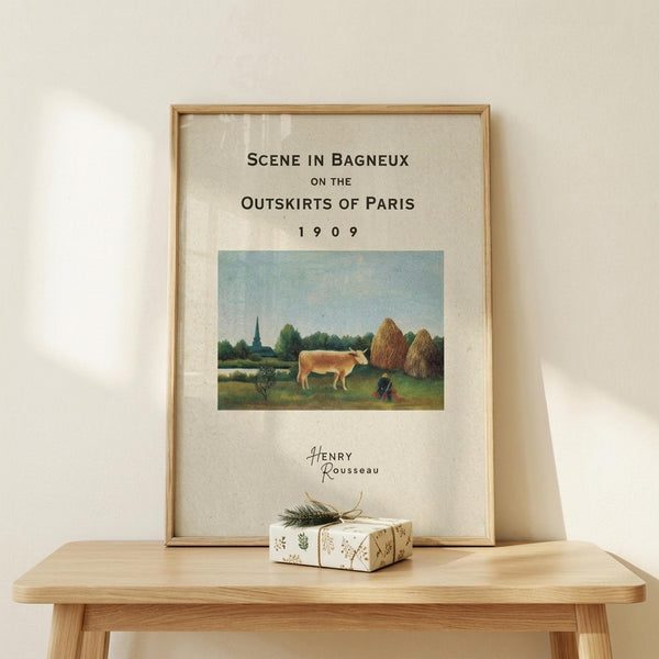 May include: Framed art print displaying a pastoral scene with a cow grazing in green countryside, haystacks, and distant church spire under blue sky. Cream matted poster in light wood frame sits on wooden bench beside small wrapped gift with botanical pattern and twine bow against white wall.