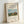 May include: Framed poster print featuring Henri Rousseau's pastoral painting of a rural French countryside scene with a brown cow, hay stacks, church spire, and figure in a field under blue sky. The cream-colored print displays black text reading "Scene in Bagneux on the Outskirts of Paris 1909" above the artwork and "Henry Rousseau" signature below. Light wood frame leans against white wall, showing realistic room display mockup with natural lighting and shadows.