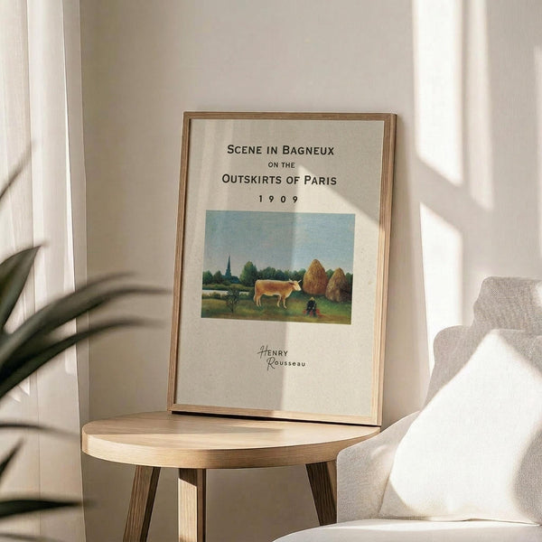 May include: Framed art print displaying a pastoral landscape with a cow, haystacks, and distant church spire under blue sky. The cream-colored poster features black text reading "Scene in Bagneux on the Outskirts of Paris 1909" and "Henry Rousseau" in script. Print sits in thin wooden frame on round wooden side table next to beige upholstered chair with throw pillow. Soft natural lighting creates gentle shadows on white wall, with green plant leaves visible in corner.