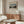 May include: Framed art print of Henri Rousseau's Eiffel Tower painting displayed above a walnut credenza in a minimalist living room. The artwork shows a twilight river scene with the Eiffel Tower visible in the distance, surrounded by trees and featuring a pale blue sky. The print has cream matting in a thin wooden frame. Room features white textured chair, glass oval coffee table, modern white lamp, and beige curtains by a window.
