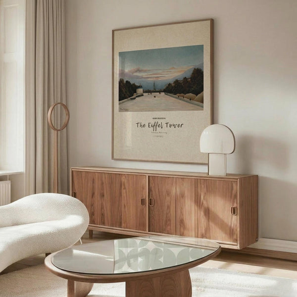 May include: Framed art print of Henri Rousseau's Eiffel Tower painting displayed above a walnut credenza in a minimalist living room. The artwork shows a twilight river scene with the Eiffel Tower visible in the distance, surrounded by trees and featuring a pale blue sky. The print has cream matting in a thin wooden frame. Room features white textured chair, glass oval coffee table, modern white lamp, and beige curtains by a window.