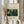 May include: Hands holding a framed art print featuring a jungle scene with lush green foliage, yellow flowers, and a small lion resting among tropical plants. The vintage-style poster has cream matting with text identifying it as a 1907 work by Henri Rousseau from the MET Museum. Natural pampas grass stems visible beside the light wood frame against soft neutral curtains and clothing.