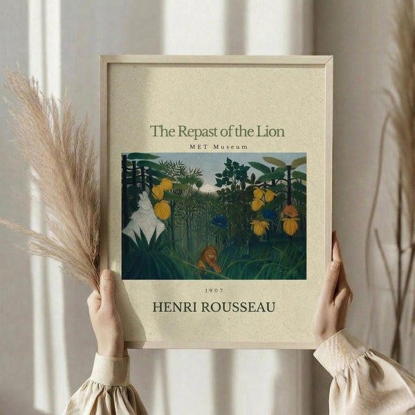 May include: Hands holding a framed art print featuring a jungle scene with lush green foliage, yellow flowers, and a small lion resting among tropical plants. The vintage-style poster has cream matting with text identifying it as a 1907 work by Henri Rousseau from the MET Museum. Natural pampas grass stems visible beside the light wood frame against soft neutral curtains and clothing.