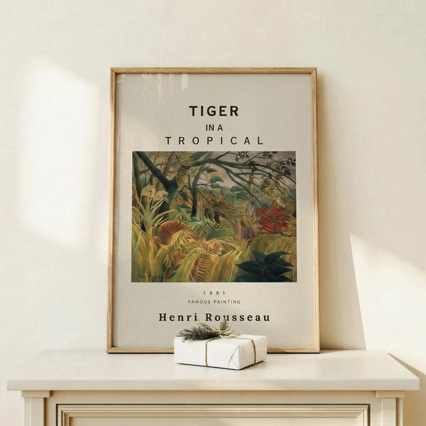 May include: Framed art print featuring Henri Rousseau's 1891 tropical jungle painting with a tiger among lush green foliage and vibrant plants. The poster has white background with black text identifying the artwork and artist. Natural wood frame sits on light wooden surface next to small white wrapped gift with twine and evergreen sprig. Soft lighting creates gentle shadows on neutral beige wall.