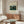 May include: Henri Rousseau jungle scene poster displayed above wooden credenza in modern living room. The vintage art print shows lush tropical foliage with yellow flowers and a resting lion, framed in thin wood frame against cream wall. Room features curved white chair, glass-top coffee table, and contemporary table lamp with dome shade.