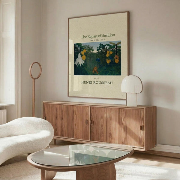 May include: Henri Rousseau jungle scene poster displayed above wooden credenza in modern living room. The vintage art print shows lush tropical foliage with yellow flowers and a resting lion, framed in thin wood frame against cream wall. Room features curved white chair, glass-top coffee table, and contemporary table lamp with dome shade.
