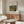 May include: Framed art print hanging above wooden sideboard in modern living room. Print features tropical jungle scene with lush green foliage and plants in painterly style, with "TIGER IN A TROPICAL" text at top and "Henri Rousseau" at bottom. Room includes white curved chair, glass oval coffee table, floor lamp, and table lamp on credenza against neutral walls.