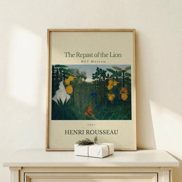 May include: Framed art print featuring Henri Rousseau's jungle scene with a lion resting among lush green tropical foliage, yellow flowers, and dense vegetation. The poster has cream-colored borders with museum-style text and is displayed in a natural wood frame on a light wooden surface next to a small wrapped gift with twine and greenery.
