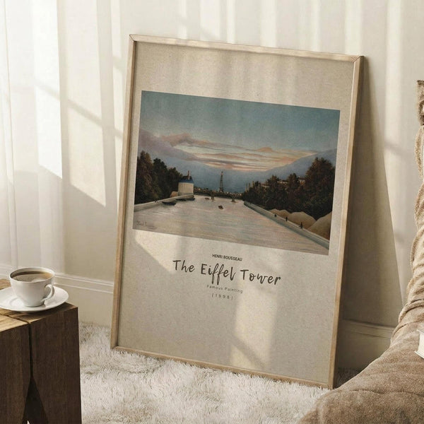 May include: Framed art print leaning against white wall corner on white textured rug, showing landscape painting with river, trees, and distant tower under cloudy sky. Wooden frame with beige matting, neutral tones throughout. Small wooden side table with white coffee cup visible on left. Cozy bedroom setting with natural lighting casting soft shadows.