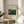 May include: Framed museum poster featuring Henri Rousseau's jungle painting with a lion among lush green foliage and yellow fruit, displayed on a round wooden side table next to a beige upholstered chair. Soft natural lighting creates gentle shadows across the neutral-toned room setting with a plant visible in the corner.