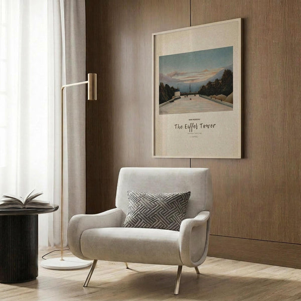 May include: Framed vintage art print displayed on wood-paneled wall showing a roadway leading to the Eiffel Tower with trees on both sides under a dusky sky. Print has cream matting with cursive text below the image. Scene includes beige upholstered armchair with geometric patterned pillow, brass floor lamp with white circular base, and dark round side table with open book.