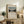 May include: Framed art print showing a tree-lined pathway leading to the Eiffel Tower under a soft blue and pink sky, displayed on a rustic wooden console alongside fern plant, stacked books, ceramic vase and table lamp in a warm, neutral-toned living room setting.
