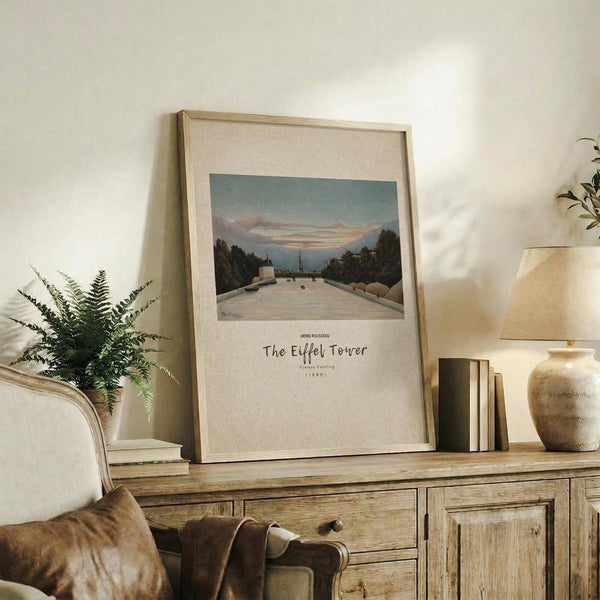May include: Framed art print showing a tree-lined pathway leading to the Eiffel Tower under a soft blue and pink sky, displayed on a rustic wooden console alongside fern plant, stacked books, ceramic vase and table lamp in a warm, neutral-toned living room setting.