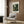 May include: Framed poster of tropical jungle scene with lush green foliage, palm fronds, and exotic plants displayed on wood-paneled wall above beige upholstered armchair with geometric patterned pillow. Modern brass floor lamp and small round table with open book complete the warm, contemporary reading nook setting.
