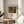 May include: Framed art print of Henri Rousseau's tropical jungle painting featuring a tiger among lush green foliage and plants during a storm. The poster has cream matting with black text below the colorful artwork. Displayed on a round wooden side table next to a beige upholstered chair with natural lighting creating soft shadows on white walls.