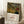 May include: Framed art print leaning against white wall showing dense jungle scene with lush green foliage, tropical plants, and hidden tiger among the vegetation. Natural wood frame with white matting displays the title and artist name below the colorful botanical artwork. Scene includes wooden side table with white coffee cup, cream textured rug, and soft natural lighting creating window shadows on the wall.