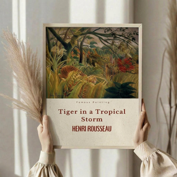 May include: Person in cream sweater holding framed art print against white curtained window, with dried pampas grass beside them. The vintage-style poster features Henri Rousseau's jungle painting with a tiger among lush green tropical foliage in golden and earth tones, displayed in thin light wood frame with cream text below the artwork.