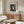 May include: Framed art print of Henri Rousseau's naive-style painting showing a wedding group portrait with bride in white dress, family members in dark clothing, and black dog, set in a forest scene. Displayed on wooden credenza in modern living room with white curved chair, glass coffee table, and cream walls.