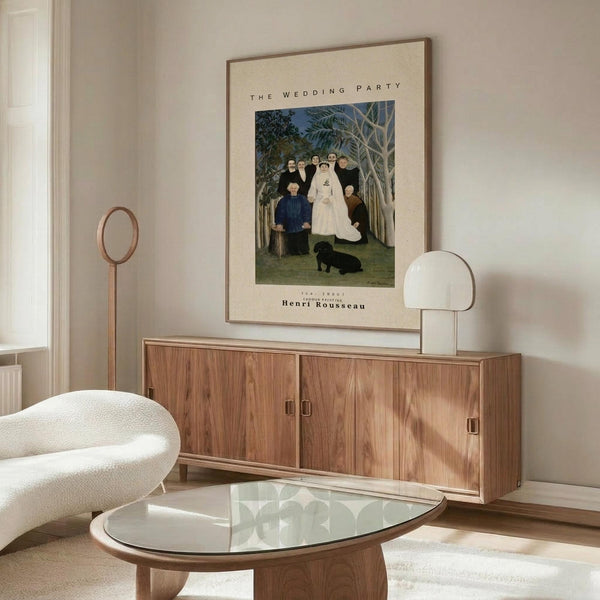 May include: Framed art print of Henri Rousseau's naive-style painting showing a wedding group portrait with bride in white dress, family members in dark clothing, and black dog, set in a forest scene. Displayed on wooden credenza in modern living room with white curved chair, glass coffee table, and cream walls.