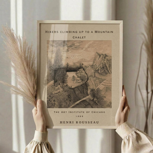 May include: Person holding framed vintage botanical print showing mountain landscape with hikers climbing rocky terrain. Sepia-toned artwork features detailed etching of cliffs, trees, and small figures on winding path. Light wooden frame held against white curtained window, dried pampas grass visible on left. Person wearing cream colored ruffled sleeve top.