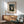 May include: Framed art print of Henri Rousseau's "The Wedding Party" painting showing a group of people in formal attire including a bride in white dress, set in a tropical forest scene with palm fronds. The print hangs on a white wall above a mid-century modern wooden credenza with sliding doors. A ceramic table lamp sits on the credenza, and a sage green upholstered chair with wooden arms is positioned nearby. Natural sunlight creates shadows across the scene.