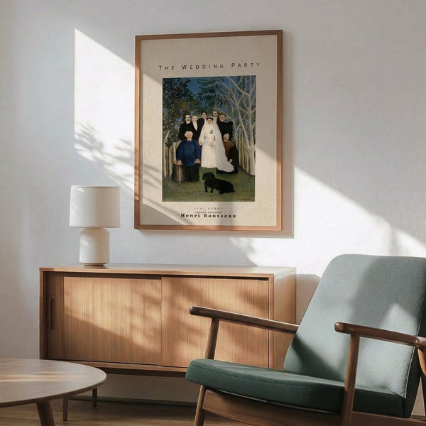 May include: Framed art print of Henri Rousseau's "The Wedding Party" painting showing a group of people in formal attire including a bride in white dress, set in a tropical forest scene with palm fronds. The print hangs on a white wall above a mid-century modern wooden credenza with sliding doors. A ceramic table lamp sits on the credenza, and a sage green upholstered chair with wooden arms is positioned nearby. Natural sunlight creates shadows across the scene.