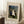 May include: Framed poster print leaning against white wall on textured cream rug, featuring Henri Rousseau's primitive-style painting of wedding party with bride in white dress, groom, and family members surrounded by lush jungle foliage with small black dog at bottom. Wooden coffee cup and saucer sit nearby on wooden block, with beige throw blanket visible at edge.