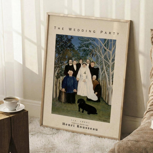 May include: Framed poster print leaning against white wall on textured cream rug, featuring Henri Rousseau's primitive-style painting of wedding party with bride in white dress, groom, and family members surrounded by lush jungle foliage with small black dog at bottom. Wooden coffee cup and saucer sit nearby on wooden block, with beige throw blanket visible at edge.