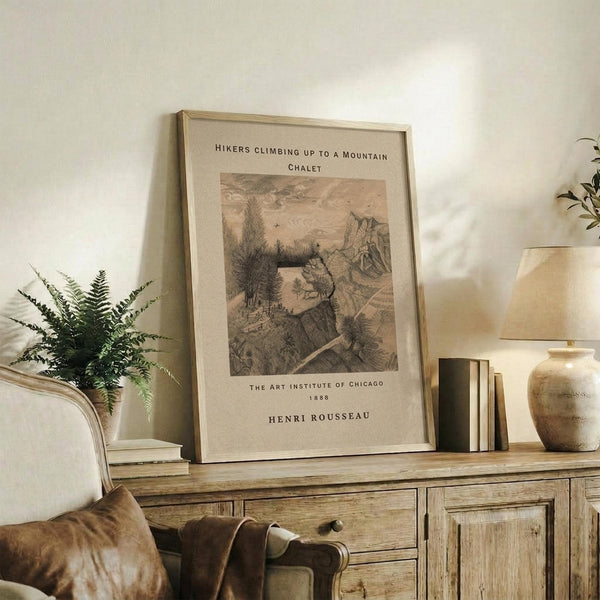 May include: Framed vintage art print leaning against white wall on rustic wooden console table. Sepia-toned illustration shows hikers climbing rocky mountain path toward chalet. Natural lighting illuminates cream-colored room with fern plant, ceramic vase, books, and beige upholstered chair. Warm, earthy home decor styling with neutral tones and organic textures.