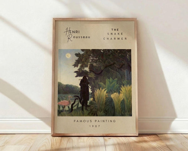 May include: Framed art print in light wood frame leaning against white wall on wooden floor. Print features Henri Rousseau's jungle scene with silhouetted figure playing flute among lush tropical plants, pink flamingo, and full moon. Beige background with black text reading "Henri Rousseau," "The Snake Charmer," "Famous Painting," and "1907." Natural lighting creates soft shadows on white fabric backdrop.