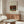 May include: Framed art print displayed above wooden sideboard in minimalist living room setting. Print shows cream background with painting of gray rabbit beside vegetables and brick wall. Room features white upholstered chair, glass oval coffee table, brass floor lamp, and neutral wall colors with natural lighting from window.