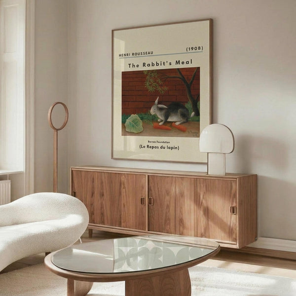 May include: Framed art print displayed above wooden sideboard in minimalist living room setting. Print shows cream background with painting of gray rabbit beside vegetables and brick wall. Room features white upholstered chair, glass oval coffee table, brass floor lamp, and neutral wall colors with natural lighting from window.